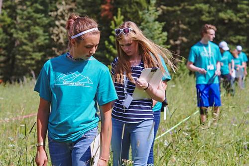 A line of youth walk outside in a field while looking down to make observations. 
